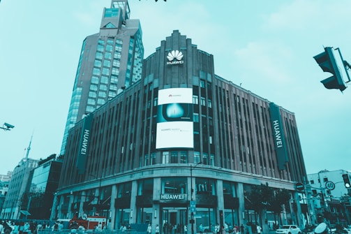 A large commercial building prominently displays the Huawei logo and name. The structure features modern architecture with vertical lines and a large digital screen advertising Huawei products. Several other high-rise buildings are visible in the background, and there is a busy street scene with people and traffic at the bottom of the image.
