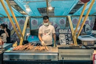 A street food vendor is standing behind a grilling station covered with skewered kebabs. The vendor is wearing a mask and gloves, indicating a focus on hygiene. The stall is decorated with bright, colorful banners featuring text and cartoon graphics. Another person is visible in the background, also wearing a mask and attending to something on a table.