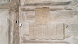 An aerial view of a construction site with large, leveled dirt surfaces marked by tire tracks. Several vehicles and a trailer are parked on the dirt path to the left, while a bulldozer is visible working on the right.