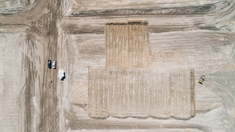 An aerial view of a construction site with large, leveled dirt surfaces marked by tire tracks. Several vehicles and a trailer are parked on the dirt path to the left, while a bulldozer is visible working on the right.