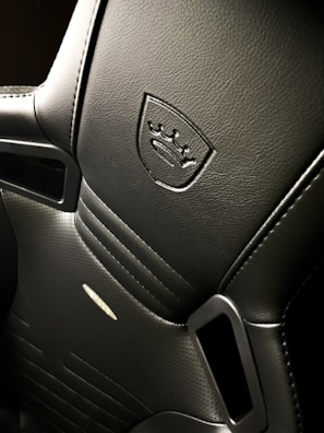 A close-up view of a sleek, black car seat with intricate stitching and a luxurious texture. An embossed logo featuring a shield and crown is prominently displayed on the upper section. The seat has perforated leather sections for ventilation and integrated grip holes on the sides, suggesting it is designed for high-performance or sport vehicles.
