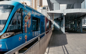 A blue tram with advertising on its side is stopped at a station platform in an urban area. The platform is covered with a metal roof, and there are several people walking in the background. Tall buildings are visible in the distance, indicating a city environment.