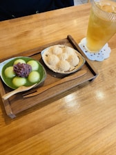 A wooden tray holds two bowls of Japanese desserts. One bowl contains green tea mochi topped with red bean paste, and the other features powder-coated mochi. A wooden spoon rests on the tray. A tall glass of iced tea is placed next to the tray on a white doily.