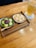 A wooden tray holds two bowls of Japanese desserts. One bowl contains green tea mochi topped with red bean paste, and the other features powder-coated mochi. A wooden spoon rests on the tray. A tall glass of iced tea is placed next to the tray on a white doily.