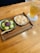 A wooden tray holds two bowls of Japanese desserts. One bowl contains green tea mochi topped with red bean paste, and the other features powder-coated mochi. A wooden spoon rests on the tray. A tall glass of iced tea is placed next to the tray on a white doily.