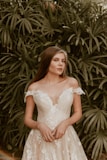 A woman in an elegant white lace dress stands in front of lush palm leaves. The dress has off-shoulder sleeves and intricate floral patterns. Her long brown hair is loose, and her expression is serene.