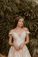 A woman in an elegant white lace dress stands in front of lush palm leaves. The dress has off-shoulder sleeves and intricate floral patterns. Her long brown hair is loose, and her expression is serene.