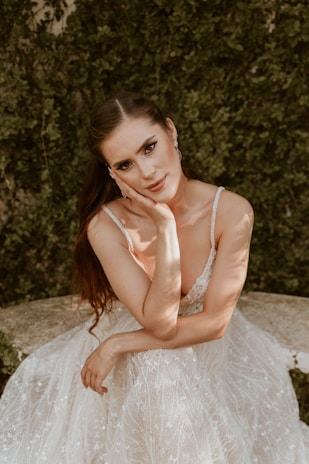 A woman with long hair wearing an elegant lace dress is seated on a stone surface. Her hand rests on her cheek as she gazes directly at the camera. The background consists of lush green foliage, and the light casts soft shadows across her face and dress.