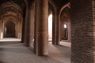 A spacious interior scene with a series of brick columns and arched ceilings. The architecture features intricate brickwork and a symmetrical design creating a sense of depth and perspective. Soft light filters through the arches, casting gentle shadows on the floor.