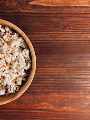 A wooden bowl filled with fluffy, freshly popped popcorn is placed on a textured wooden surface. The contrast between the light-colored popcorn and the rich, warm tones of the wood creates a cozy, inviting atmosphere.