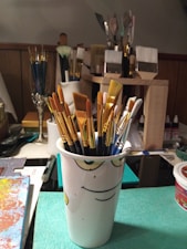 A variety of paintbrushes are grouped together in a white cup with a face design, situated on a table. Several different types of brushes, including flat, round, and fan brushes, can be seen with tips in shades of brown, white, and gold. The background consists of more brushes in containers, various art supplies, and a partially visible painting on the left.