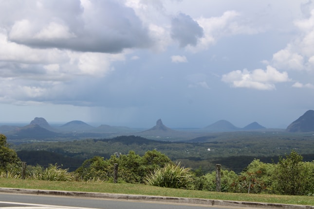 A scenic landscape featuring a range of distinctive mountain peaks against a backdrop of partly cloudy skies. The foreground includes lush green vegetation and a road lined with posts. The mountains appear in varying sizes and shapes, adding to the dramatic skyline.