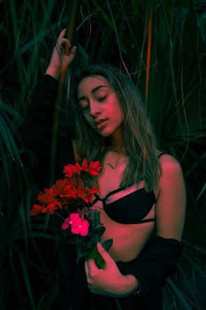A woman with eyes closed, wearing minimal clothing and surrounded by lush green foliage, holds a bouquet of vibrant red and pink flowers. The scene is dimly lit with muted natural lighting and evokes a serene and introspective mood.
