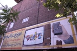 An outdoor advertising board is displayed on a brick wall with some trees partially obscuring it. The board features an illustration of a vintage television showing animated characters and a hand holding a remote control. Text in a foreign script is present on the left side of the billboard.