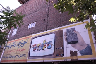 An outdoor advertising board is displayed on a brick wall with some trees partially obscuring it. The board features an illustration of a vintage television showing animated characters and a hand holding a remote control. Text in a foreign script is present on the left side of the billboard.