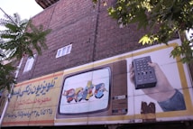 An outdoor advertising board is displayed on a brick wall with some trees partially obscuring it. The board features an illustration of a vintage television showing animated characters and a hand holding a remote control. Text in a foreign script is present on the left side of the billboard.