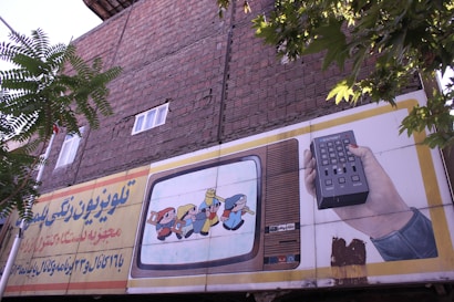 An outdoor advertising board is displayed on a brick wall with some trees partially obscuring it. The board features an illustration of a vintage television showing animated characters and a hand holding a remote control. Text in a foreign script is present on the left side of the billboard.
