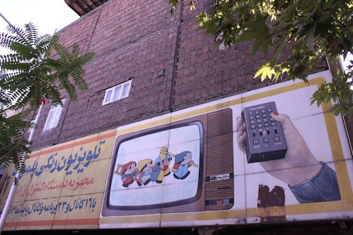 An outdoor advertising board is displayed on a brick wall with some trees partially obscuring it. The board features an illustration of a vintage television showing animated characters and a hand holding a remote control. Text in a foreign script is present on the left side of the billboard.