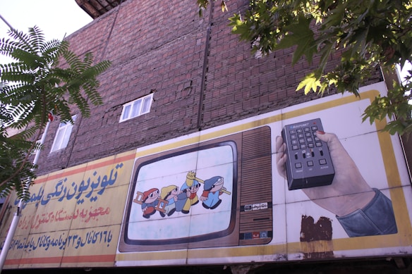 An outdoor advertising board is displayed on a brick wall with some trees partially obscuring it. The board features an illustration of a vintage television showing animated characters and a hand holding a remote control. Text in a foreign script is present on the left side of the billboard.