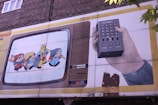 A mural on a building wall depicts a vintage television with cartoon characters inside. One character wears a yellow hat and appears to be leading others while holding hands. Next to the television, a hand holds a large remote control with various buttons. The scene has a nostalgic, retro feel.
