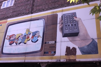 A mural on a building wall depicts a vintage television with cartoon characters inside. One character wears a yellow hat and appears to be leading others while holding hands. Next to the television, a hand holds a large remote control with various buttons. The scene has a nostalgic, retro feel.