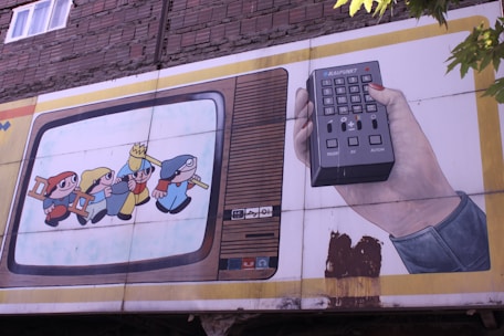 A mural on a building wall depicts a vintage television with cartoon characters inside. One character wears a yellow hat and appears to be leading others while holding hands. Next to the television, a hand holds a large remote control with various buttons. The scene has a nostalgic, retro feel.