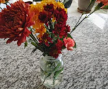 A vibrant bouquet of pink, yellow, and orange flowers in a vintage mason jar on a rustic wooden table.