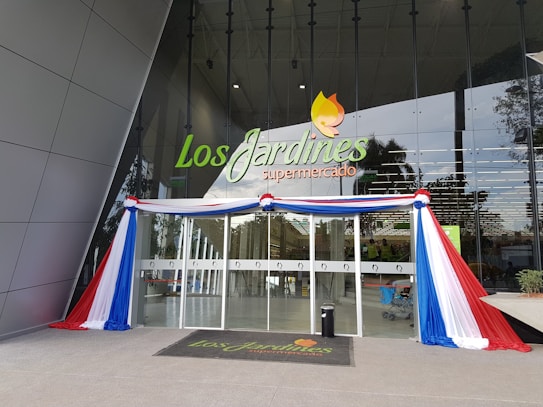 An entrance to a supermarket named 'Los Jardines' is adorned with red, white, and blue fabric draped around the glass doors. The signage above the entrance displays the supermarket's name in green and orange letters. The facade is modern with reflective glass, and a black mat with the store's logo is placed at the entrance.