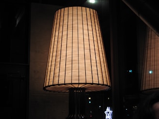 A large, elegant lampshade with a warm glow. The lampshade is woven with vertical lines and emits a soft, ambient light. It is placed in a dimly lit room with reflective glass surfaces in the background.