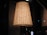 A large, elegant lampshade with a warm glow. The lampshade is woven with vertical lines and emits a soft, ambient light. It is placed in a dimly lit room with reflective glass surfaces in the background.