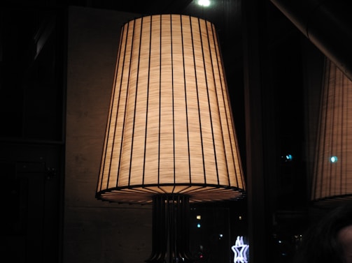 A large, elegant lampshade with a warm glow. The lampshade is woven with vertical lines and emits a soft, ambient light. It is placed in a dimly lit room with reflective glass surfaces in the background.