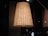 A large, elegant lampshade with a warm glow. The lampshade is woven with vertical lines and emits a soft, ambient light. It is placed in a dimly lit room with reflective glass surfaces in the background.
