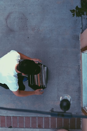 A person crouching on a concrete surface while sorting through a crate filled with vinyl records. The individual is wearing a white shirt and a black cap. The scene is viewed from above, capturing part of the sidewalk and a brick edge.