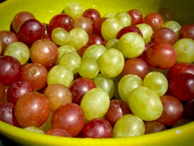 A close-up view of a bowl filled with a mix of red and green grapes. The grapes appear fresh and juicy, their skins glistening under the light. They are nestled in a bright yellow bowl, enhancing the vibrancy of the scene.