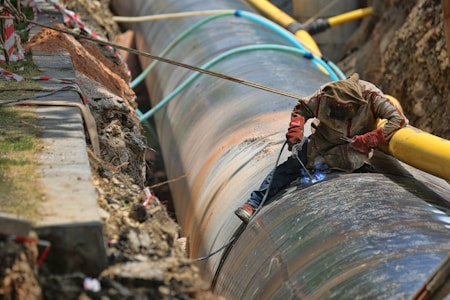 A worker is engaged in welding a large metal pipeline. The pipeline is situated in a trench lined with earth and concrete. The worker is wearing protective gear, including a hood and gloves, and is using welding equipment that emits a bright blue light. Various cables and pipes are positioned around the trench.