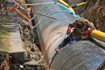 A worker is engaged in welding a large metal pipeline. The pipeline is situated in a trench lined with earth and concrete. The worker is wearing protective gear, including a hood and gloves, and is using welding equipment that emits a bright blue light. Various cables and pipes are positioned around the trench.