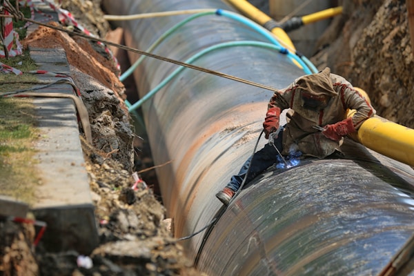 A worker is engaged in welding a large metal pipeline. The pipeline is situated in a trench lined with earth and concrete. The worker is wearing protective gear, including a hood and gloves, and is using welding equipment that emits a bright blue light. Various cables and pipes are positioned around the trench.