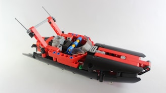 A LEGO model resembling a vehicle with a red and black color scheme. It includes various small components like gears and axles, with two antenna-like extensions at the back and a transparent windshield at the front.