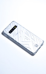 A smartphone with a transparent, geometric-patterned case is lying on a white surface. The case covers the back and sides of the phone, revealing its camera lenses and branding. The overall aesthetic is sleek and modern.