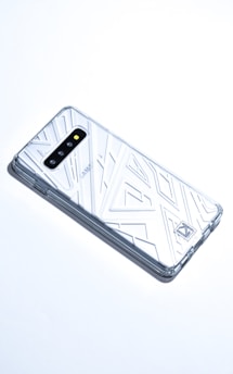 A smartphone with a transparent, geometric-patterned case is lying on a white surface. The case covers the back and sides of the phone, revealing its camera lenses and branding. The overall aesthetic is sleek and modern.