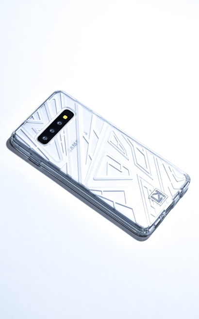 A smartphone with a transparent, geometric-patterned case is lying on a white surface. The case covers the back and sides of the phone, revealing its camera lenses and branding. The overall aesthetic is sleek and modern.