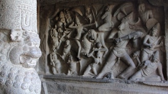 Ancient stone carving depicting a mythological scene with numerous figures engaged in battle, featuring a prominent figure with a bull-like head holding a weapon. The relief is intricately detailed, capturing dynamic postures and expressive gestures. A carved pillar with elaborate design stands on the left.