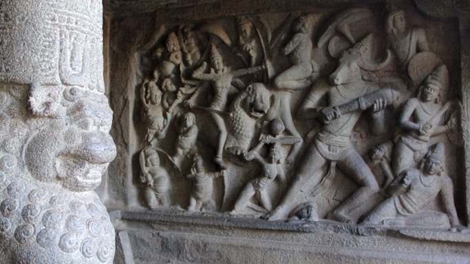 Ancient stone carving depicting a mythological scene with numerous figures engaged in battle, featuring a prominent figure with a bull-like head holding a weapon. The relief is intricately detailed, capturing dynamic postures and expressive gestures. A carved pillar with elaborate design stands on the left.