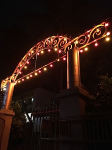 An archway adorned with decorative, swirling metalwork is illuminated by a string of colorful lights, creating a festive atmosphere. The lights are mainly red, yellow, and orange, casting a warm glow against the dark background. The structure beneath the arch appears to be a gate with pointed tips and some surrounding greenery, while a building can be seen in the shadows.