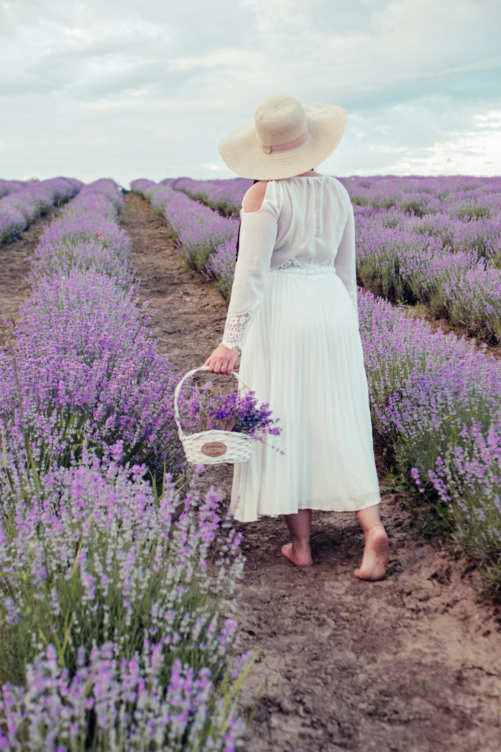 昼間、紫色の花畑を歩く籠を持った白いドレスを着た女性