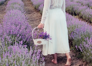woman in white dress holding basket walking on purple flower field during daytime