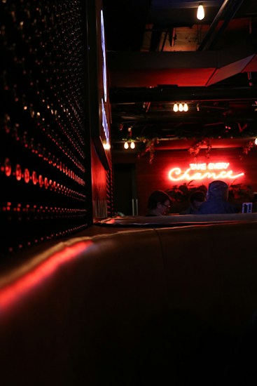 A dimly lit bar or lounge setting featuring neon signs, with a cozy and intimate atmosphere. The scene includes subtle red and orange lighting reflecting off surfaces. Patrons can be seen in the background, sitting at a table.
