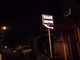 A street scene at night featuring a brightly lit sign reading 'Cafe Britos'. The sign is prominently displayed, standing on a tall pole with neon lights. Several people are walking along the street, and a parked vehicle is visible in the background. The atmosphere is dark with a few streetlights illuminating the surroundings.