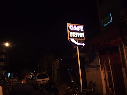 A street scene at night featuring a brightly lit sign reading 'Cafe Britos'. The sign is prominently displayed, standing on a tall pole with neon lights. Several people are walking along the street, and a parked vehicle is visible in the background. The atmosphere is dark with a few streetlights illuminating the surroundings.
