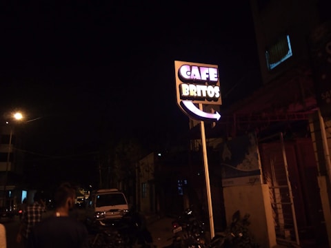 A street scene at night featuring a brightly lit sign reading 'Cafe Britos'. The sign is prominently displayed, standing on a tall pole with neon lights. Several people are walking along the street, and a parked vehicle is visible in the background. The atmosphere is dark with a few streetlights illuminating the surroundings.